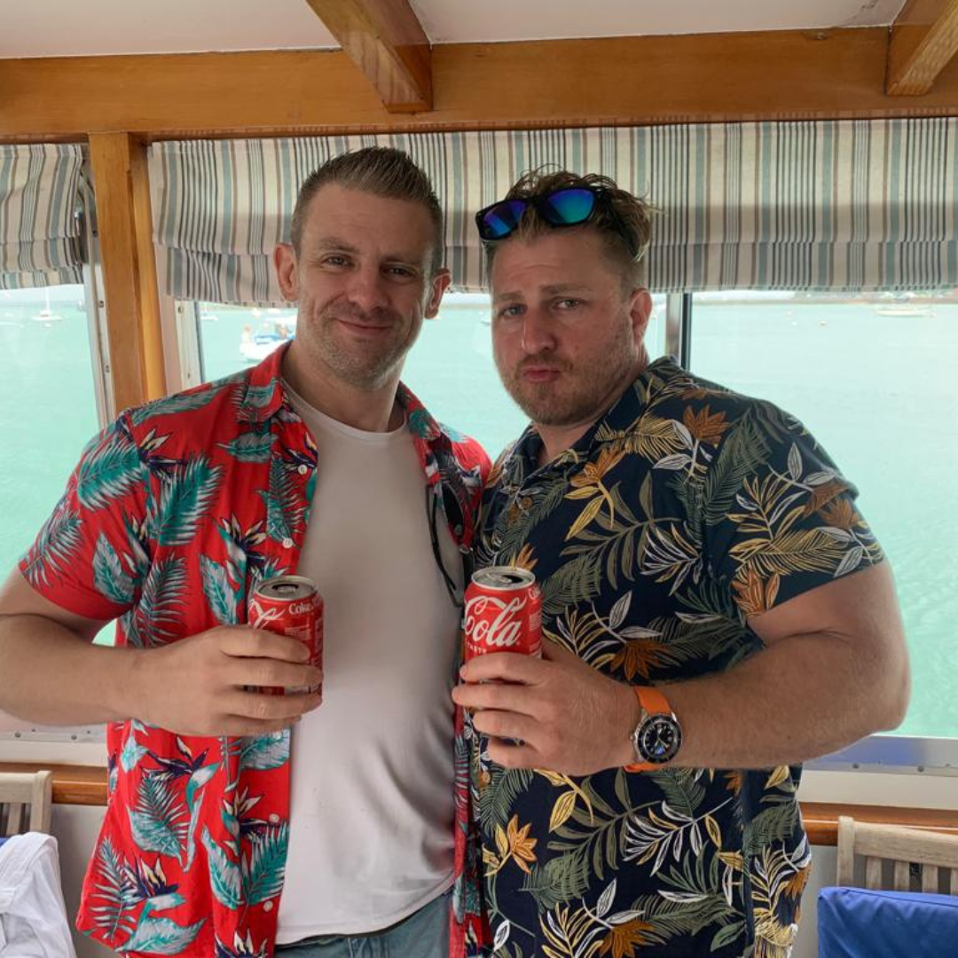 Two men smiling while sitting on a boat, one in a plaid shirt and the other in a floral shirt, with an overcast sky and ocean in the background.