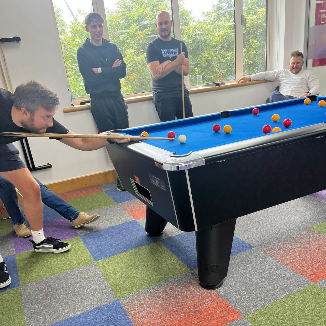 A person is playing pool, aiming at a spread of red and yellow balls on a blue pool table, while three other individuals watch.
