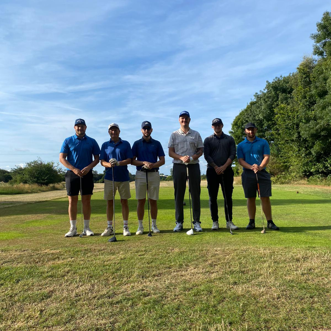 Six golfers standing in a row on a golf course, each holding a golf club, with a background of clear skies, trees, and greenery.