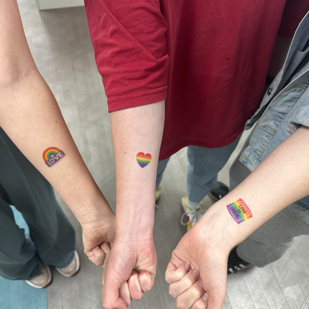 Three individuals showcasing a temporary tattoo which is LGBTQ+ flag on their wrist.