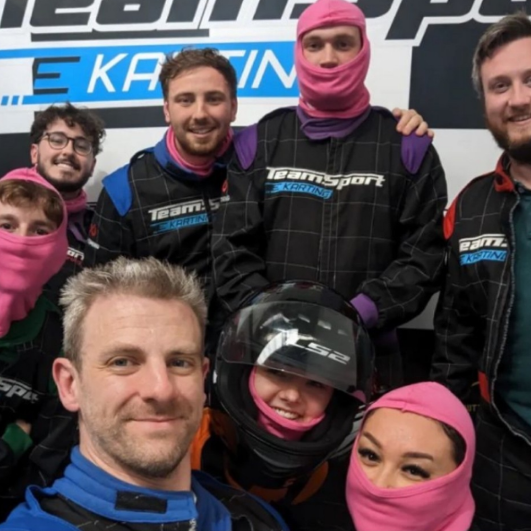 Group of people wearing go-kart racing gear, including helmets, and protective overalls, standing in front of a 'TeamSport Karting' banner.