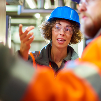 A female engineer wearing a blue hard hat, safety glasses, and an orange hi-vis jacket speaks animat