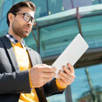 A man wearing glasses and a yellow jumper under a grey blazer reviews a tablet device outdoors in fr