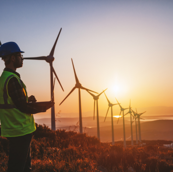 An engineer in a hi-vis vest and blue hard hat stands on a hillside at sunset, looking out over a ro