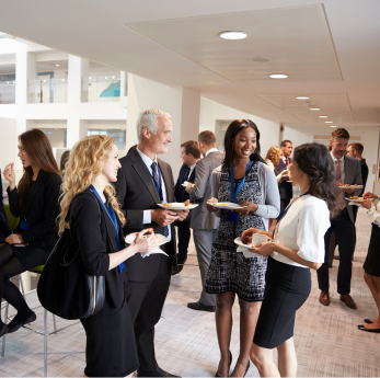 A group of professionally dressed people mingle and chat at a business networking event, holding pla